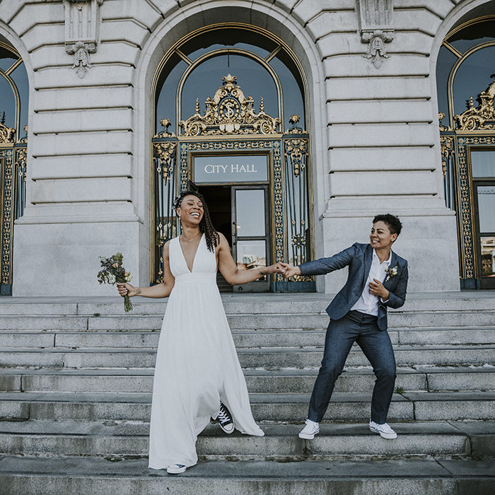 San Francisco City Hall Wedding – Two brides hold hands dancing on the grand steps in front of its elegant gold-accented doors