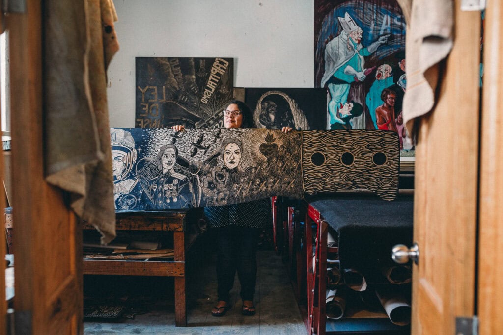  A woman holding a large piece of artwork in a studio with various paintings and creative materials, capturing a branding photoshoot in Oaxaca 