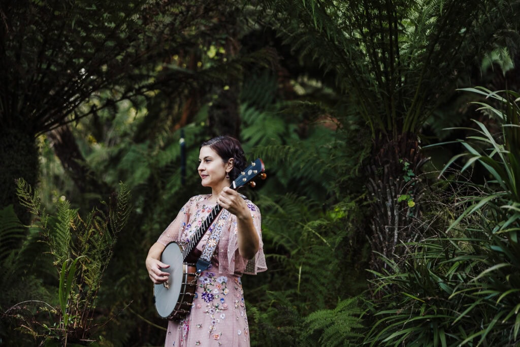 Woman playing banjo outdoors in a dense, green forest environment.