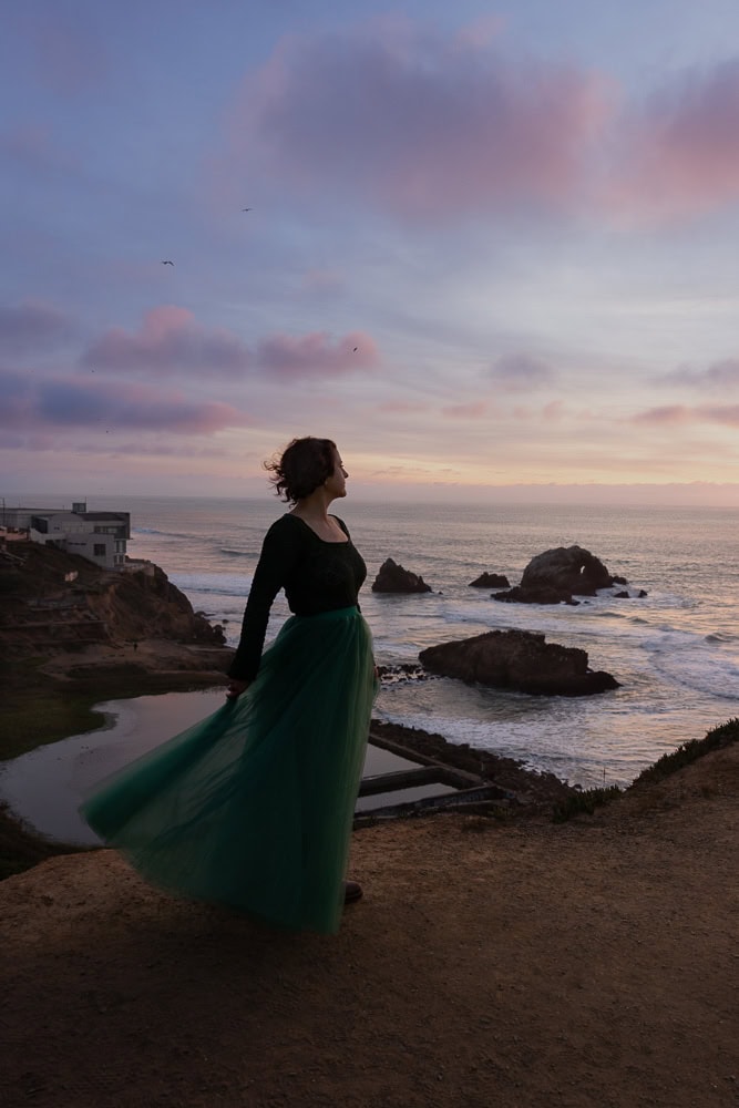 Woman in flowing green skirt overlooking coastal landscape at dusk.