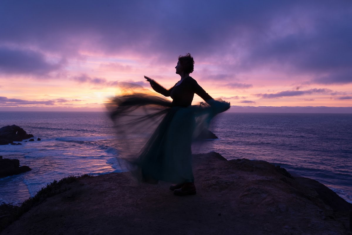Sunset silhouette of a woman on a rocky coast at dusk, with vibrant purple and orange sky.