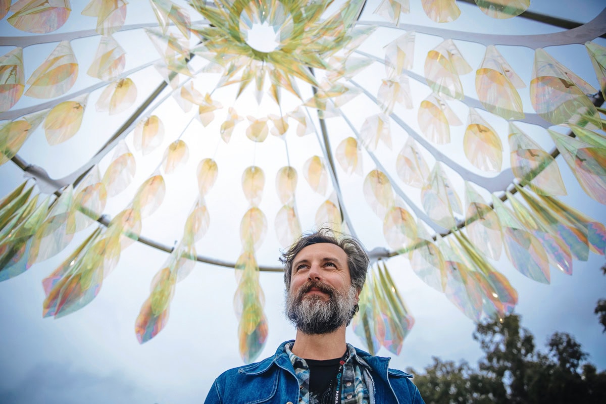Vibrant outdoor art installation featuring colorful hanging decorations and a smiling man beneath.