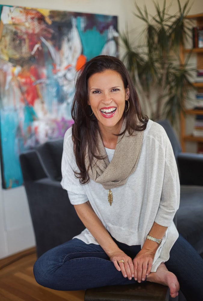 Woman smiling in a cozy, stylish home office with colorful artwork and plants.