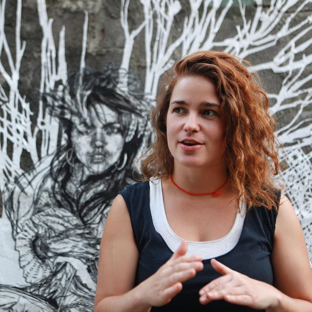 Portrait of a young woman with curly red hair in front of a black and white mural background.