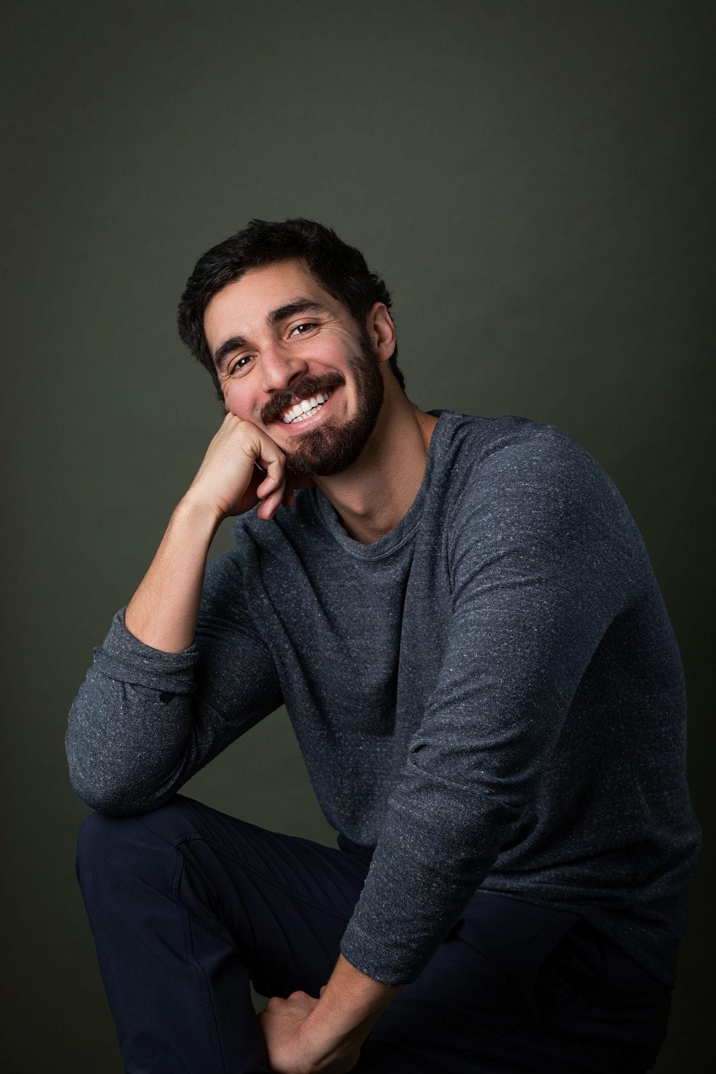 A professional portrait of a smiling man with dark hair and beard, wearing a gray sweater, against a.