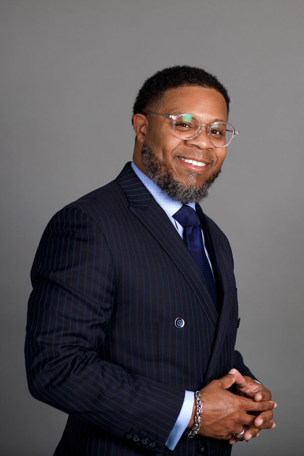 Professional portrait of a man in a navy pinstripe suit with glasses, smiling confidently.