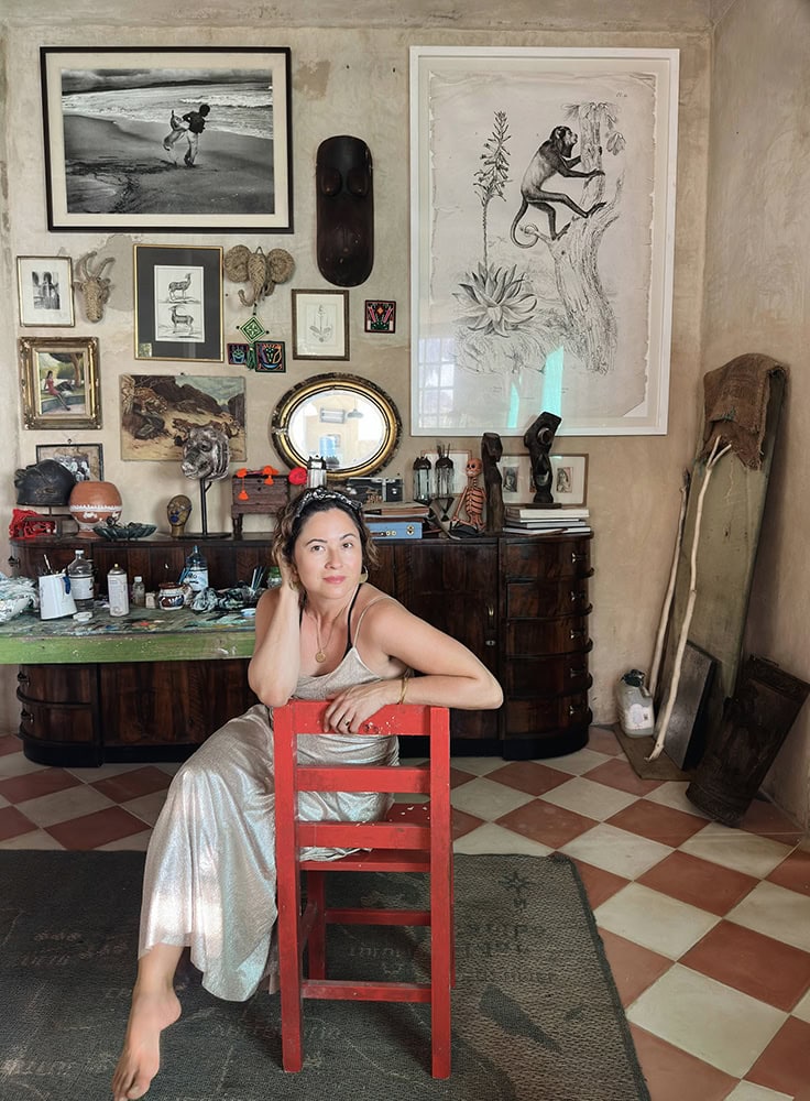Elegant woman sitting on a red chair in a room decorated with artwork and vintage furniture.
