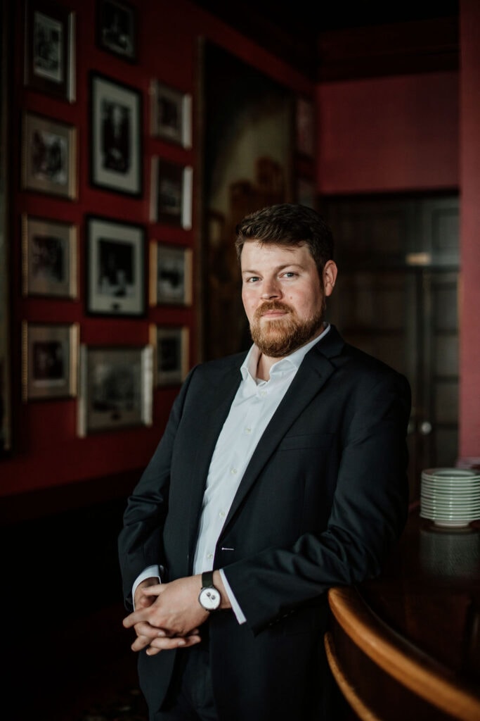 Confident businessman in a suit standing in a stylish, modern office setting with framed photos on t.