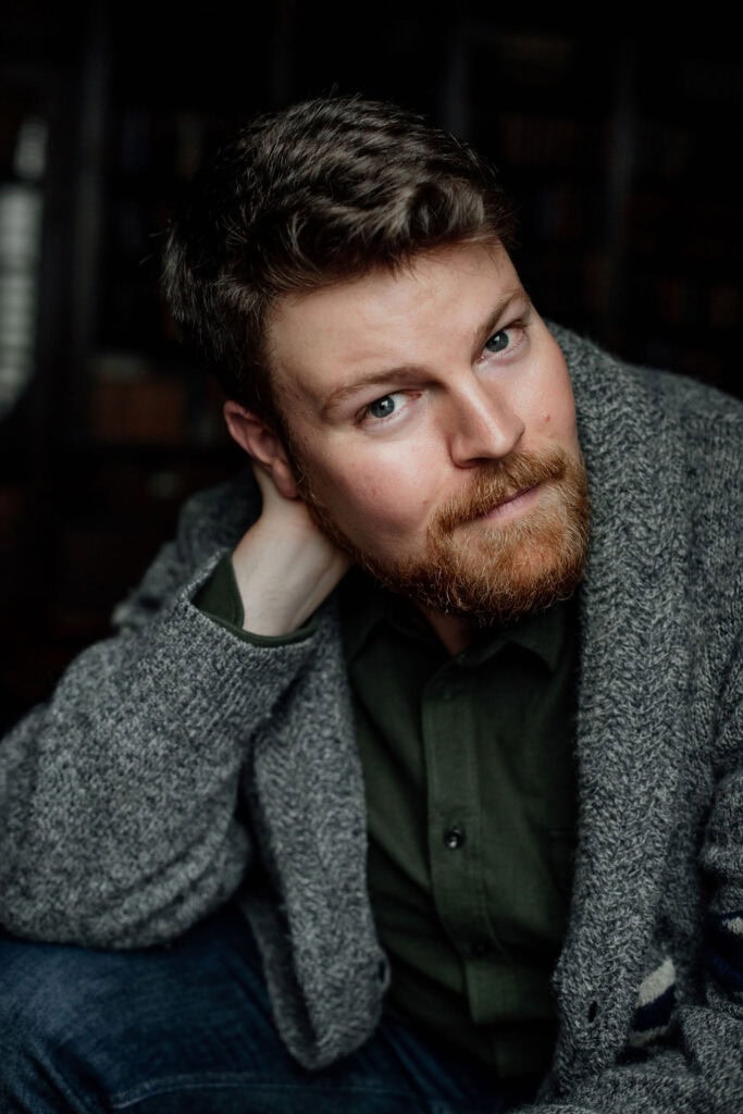 Portrait of a young man with a beard and blue eyes, wearing a gray blazer and green shirt.
